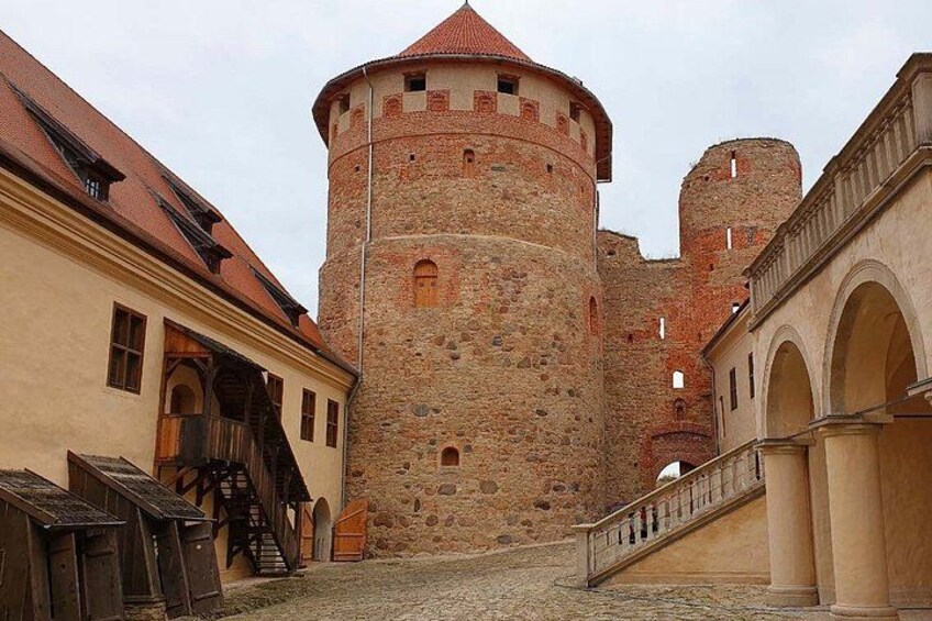 Ein historischer Steinturm mit rotem Dach, eine Kopfsteinpflasterstraße und traditionelle Gebäude mit Rundbogenfenstern. 3