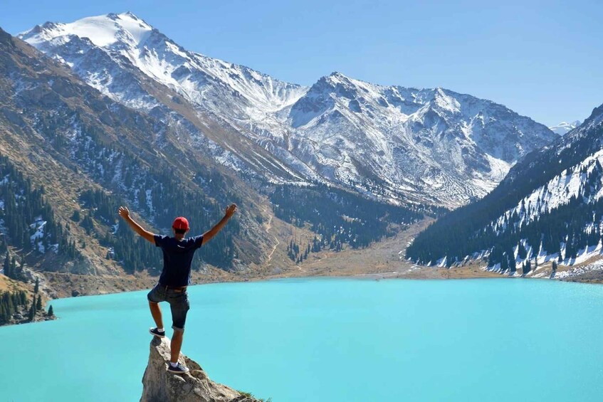 A person standing on a rock with arms raised, overlooking a turquoise lake surrounded by snow-capped mountains. 1