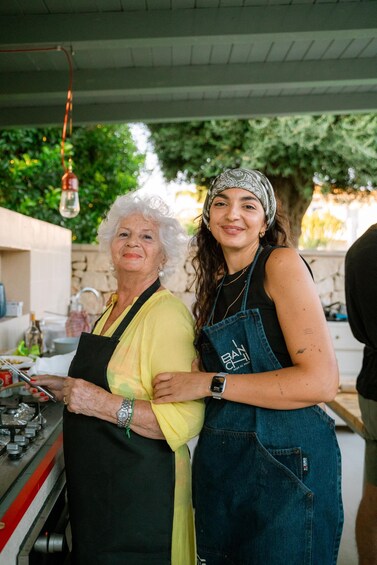 Two people standing in an outdoor cooking area with a gas stove and cooking utensils. 3