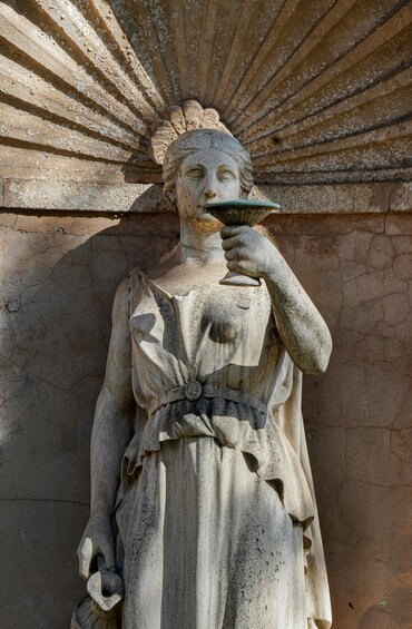 A stone statue of a woman with a fan, standing against a textured wall. 3