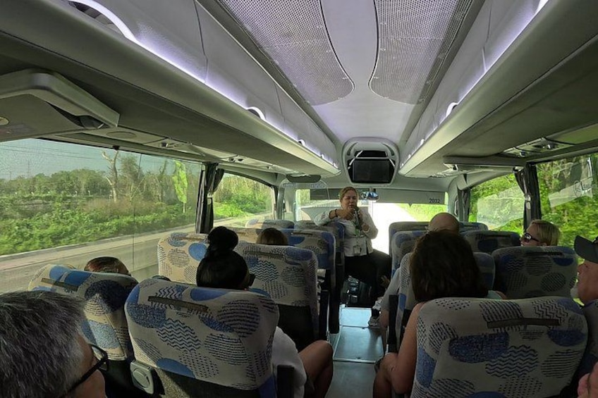 A bus interior with passengers seated, a driver at the front, and a view of greenery outside. 4