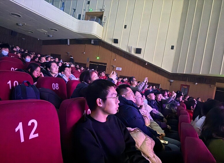 An indoor auditorium with rows of red seats, some occupied by people. 2