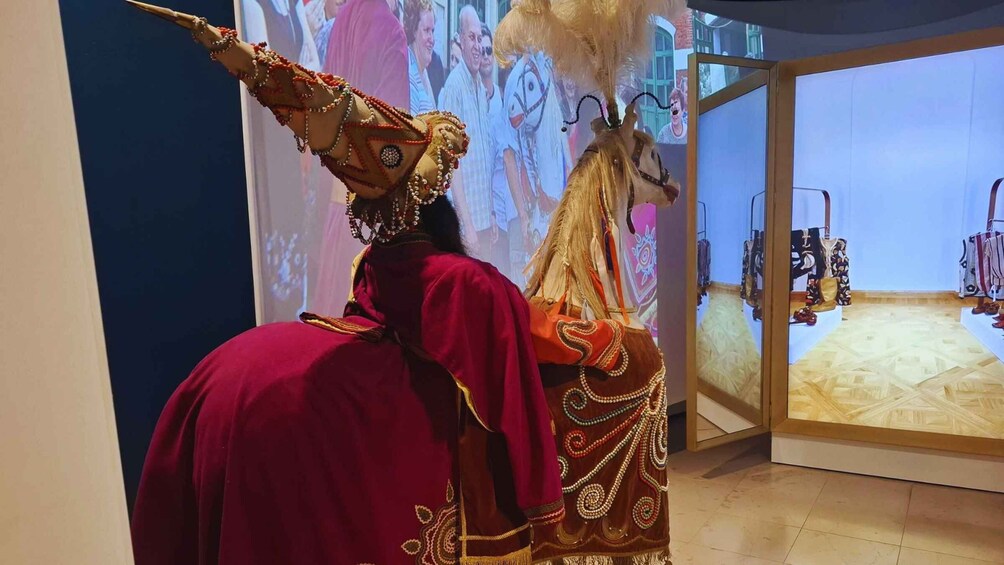 A person in traditional attire with a horse, in a room with a projection screen displaying people. 5