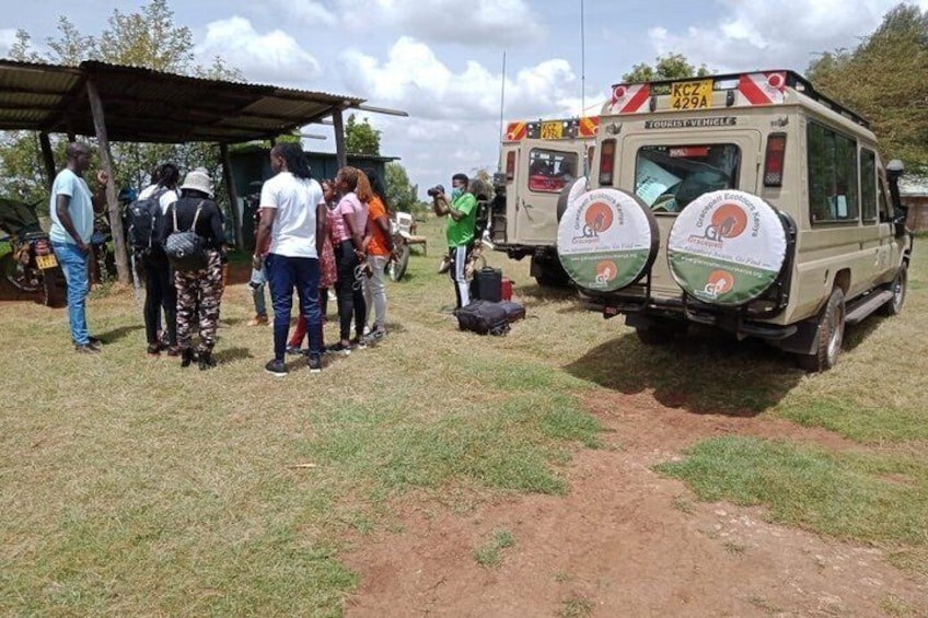 A group of people standing near a safari vehicle with a roof, and a dirt path in a grassy area. 5