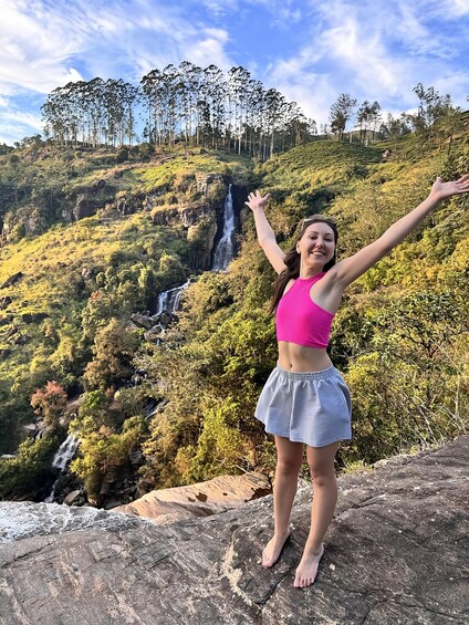 A person standing on a rock with arms raised, overlooking a waterfall and lush greenery. 2