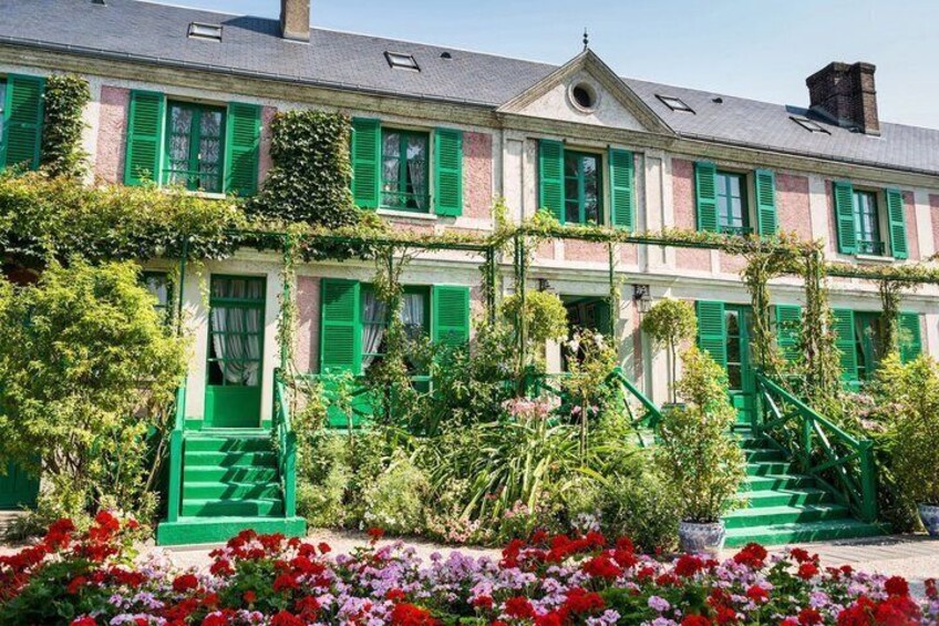 A building with green shutters and a garden with red and pink flowers. 1