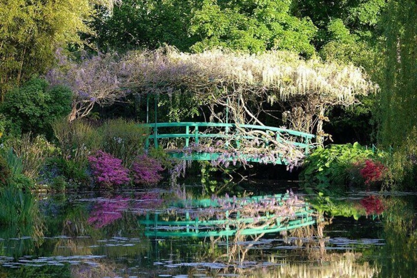 A green bridge over a pond surrounded by lush greenery and flowering plants. 2