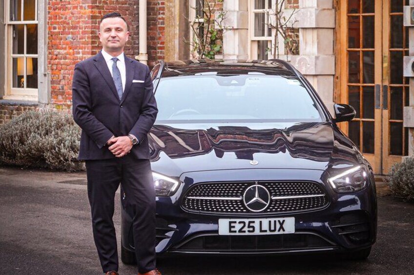 A man in a suit standing next to a black Mercedes-Benz E-Class car with a license plate reading 'E25 LUX' in front of a brick building with large windows. 2