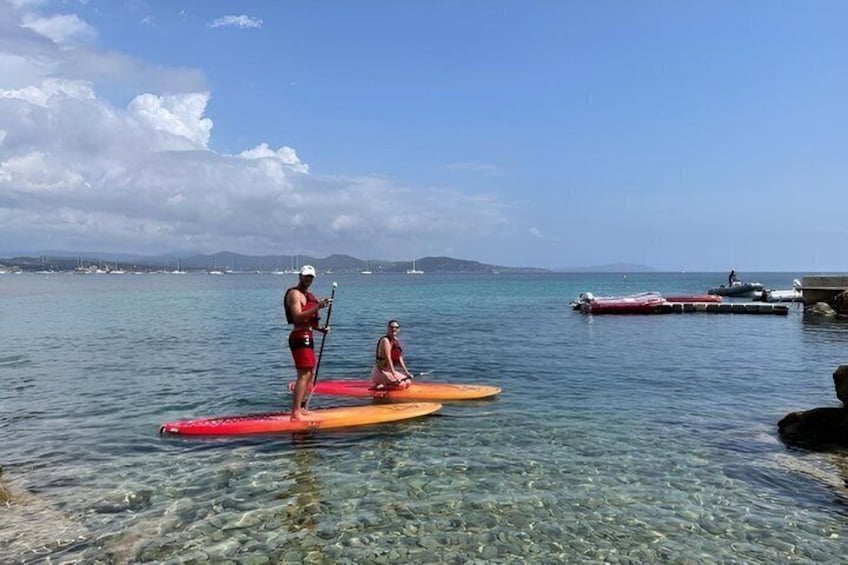 Two people paddleboarding on a calm sea with clear water, boats in the background, and a distant shoreline. 3