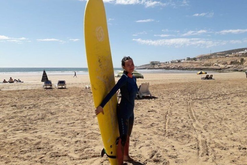 A person holding a yellow surfboard on a sandy beach with lounge chairs and umbrellas in the background. 5
