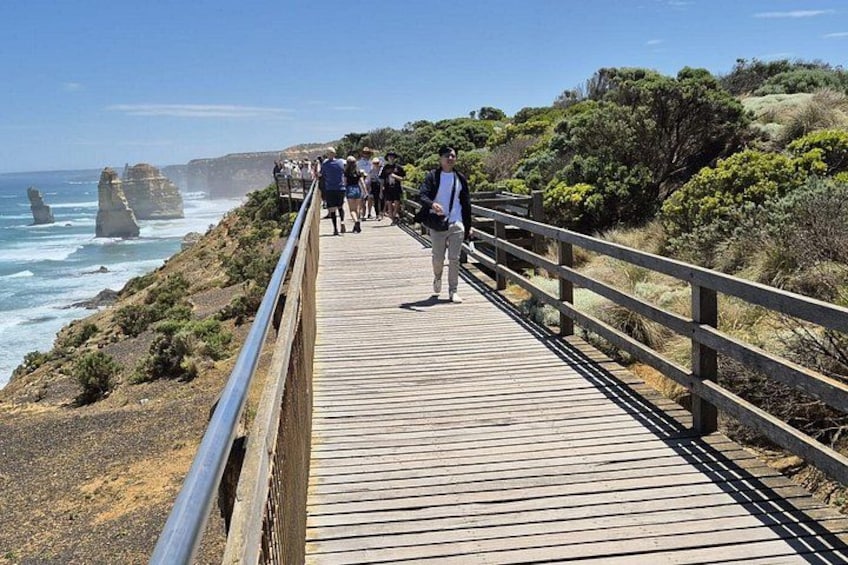 A wooden boardwalk with people walking along it, overlooking the ocean with rocky cliffs in the background. 1