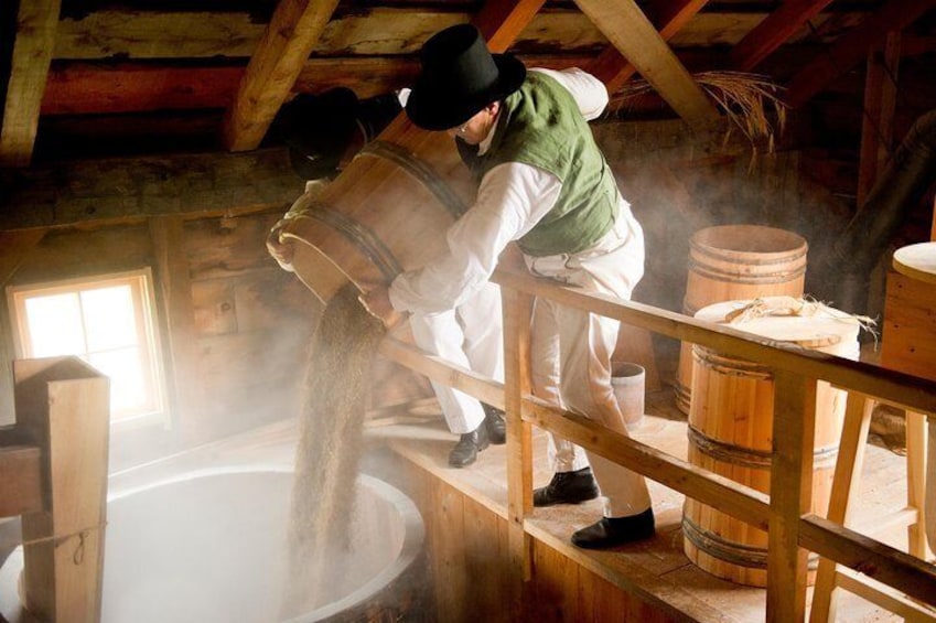 A person pouring grain into a large wooden vat in a rustic setting. 5