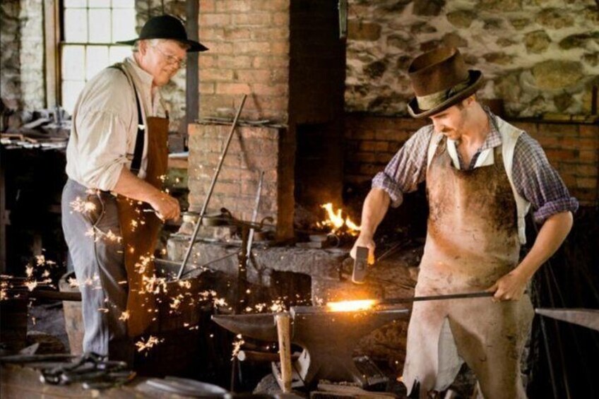 Two individuals working with a forge, sparks flying, in a brick building with a stone wall. 4