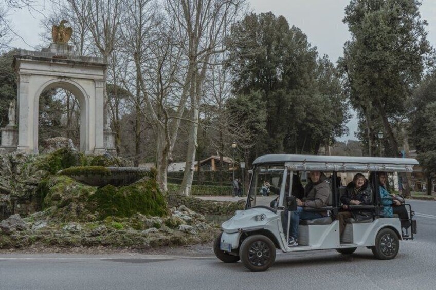 A white vehicle with passengers on a road near an archway and trees. 5