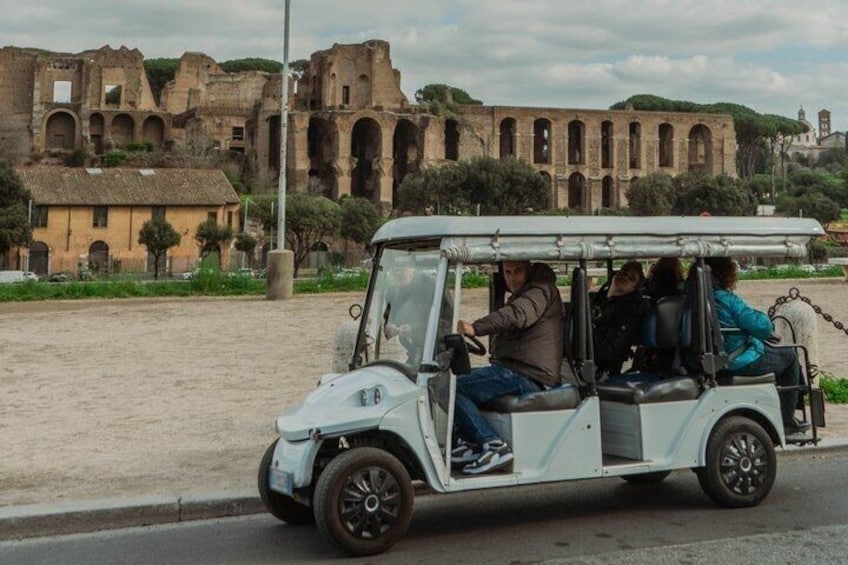 A white golf cart with passengers on a road with ancient ruins in the background. 4