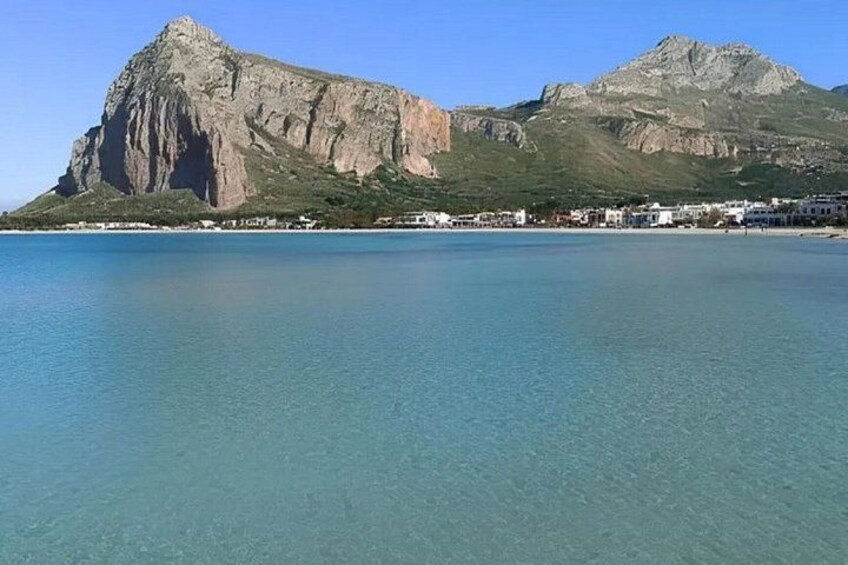 A coastal landscape with clear blue waters, a prominent rocky mountain, and a small settlement with buildings along the shore. 3