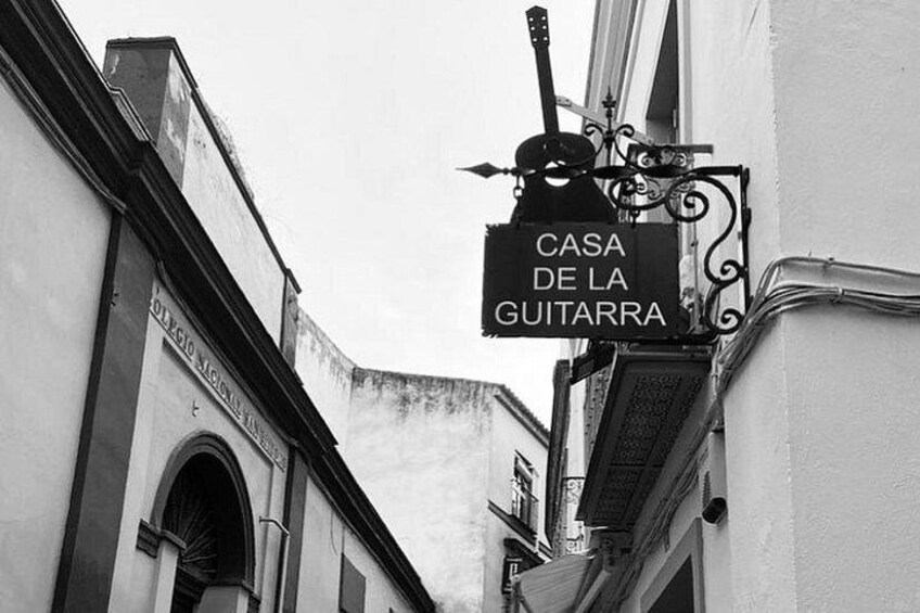 A narrow street with buildings on either side, a sign reading 'CASA DE LA GUITARRA,' and a decorative metal sign above it. 3
