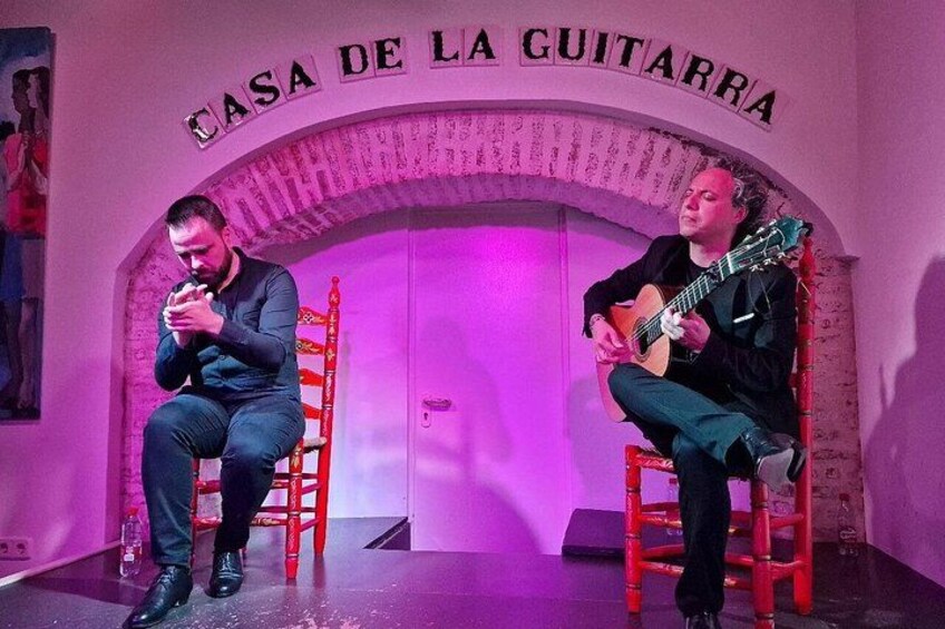 Deux personnes sont assises sur des chaises, l’une joue de la guitare, dans une pièce où est affiché un panneau indiquant « CASA DE LA GUITARRA ». 4