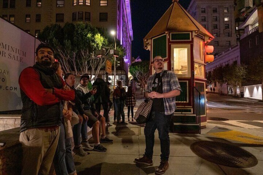 A man standing on a sidewalk at night with a group of people sitting on benches. 2