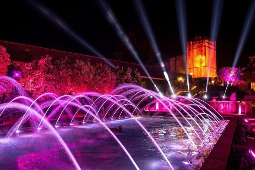 A nighttime fountain with water jets illuminated by pink and purple lights, a building with a clock tower in the background, and a tree with pink lighting. 3