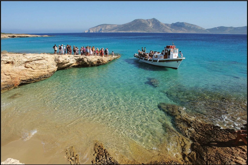 A rocky shoreline with clear turquoise water, a boat with passengers, and distant mountains. 3