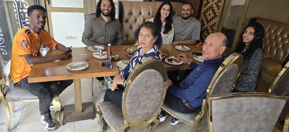 A group of people seated around a wooden table with a tufted headrest, in a room with patterned wallpaper and a brown leather couch. 5