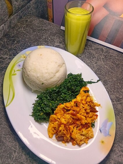 A plate with a round white food item, green leafy vegetables, and a yellow-orange dish, accompanied by a glass of yellow liquid. 4