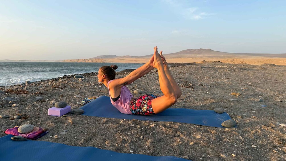 A person performing a yoga pose on a mat by a rocky beach with the ocean and mountains in the background. 4