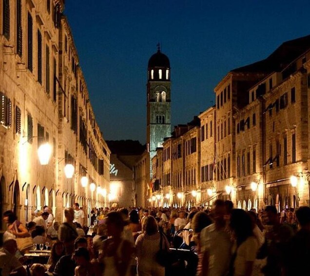 A crowded street at dusk with illuminated buildings and a prominent tower in the background. 1