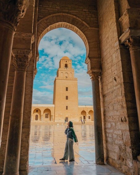 An arched corridor with a person standing in the center, looking at a tall tower with a minaret. 1