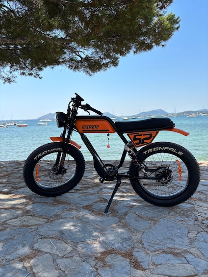 A black and orange motorcycle with 'TRIONFALLE' on the wheels, parked on a stone surface by a body of water with boats and a distant mountain. 5
