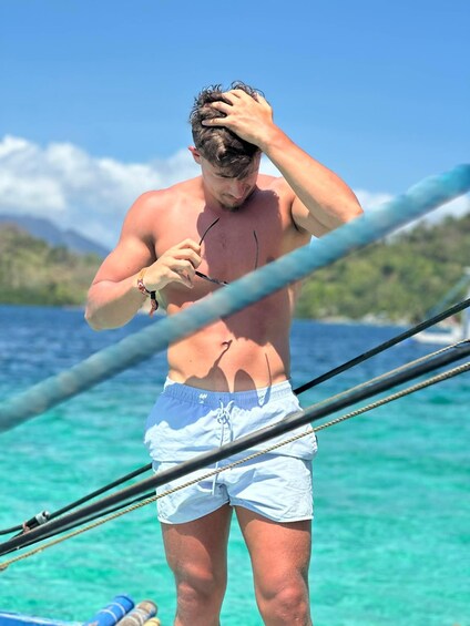 A person standing on a boat, wearing white swim shorts, with a clear blue sky and turquoise water in the background. 2