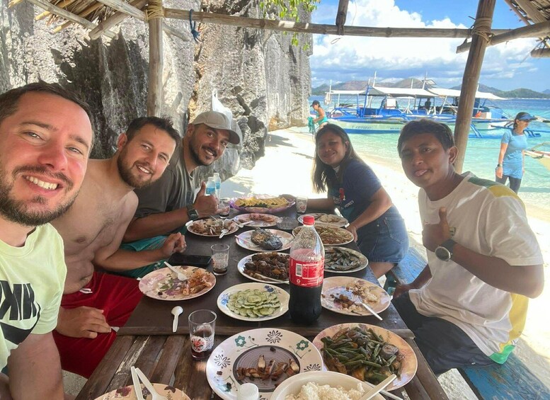 A group of people dining at a wooden table with a view of boats and a beach. 3