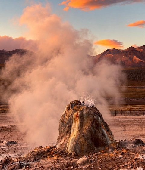 A geyser erupting with steam in a dry, rocky landscape with mountains in the background. 1