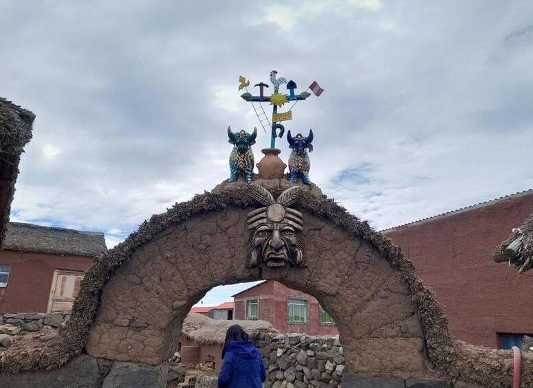 A traditional stone archway with indigenous carvings and figures, leading to a village with red-roofed buildings. 3