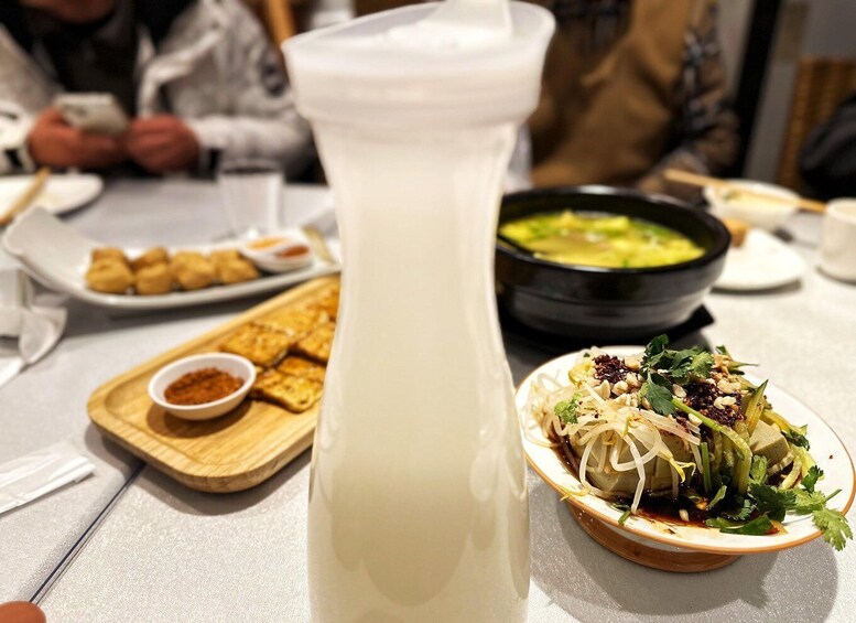 A table with a large white beverage container, a bowl of soup, a plate of fried items, and a dish with vegetables and peanuts. 5