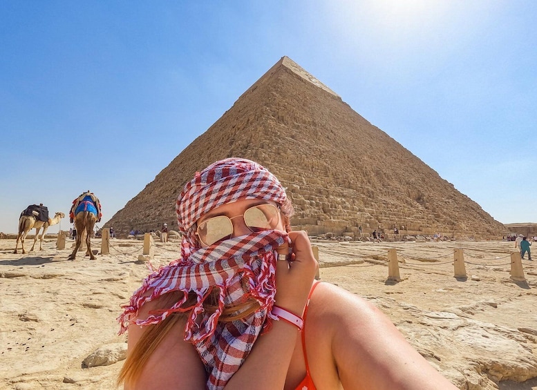 A person in a checkered headscarf and red bikini lying on the sand with a pyramid in the background. 1