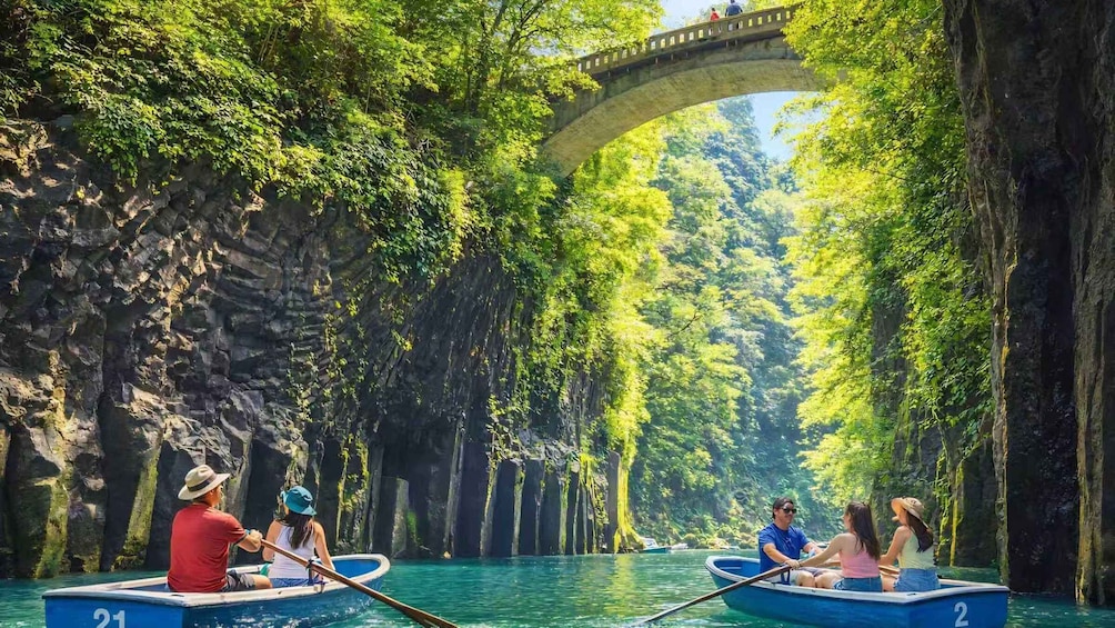 Two boats on a river with a stone bridge overhead and dense greenery on either side. 1