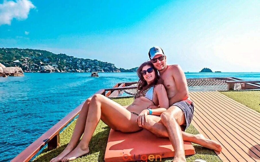 A couple relaxing on a wooden deck by a body of water with a boat in the distance and a hilly landscape in the background. 3
