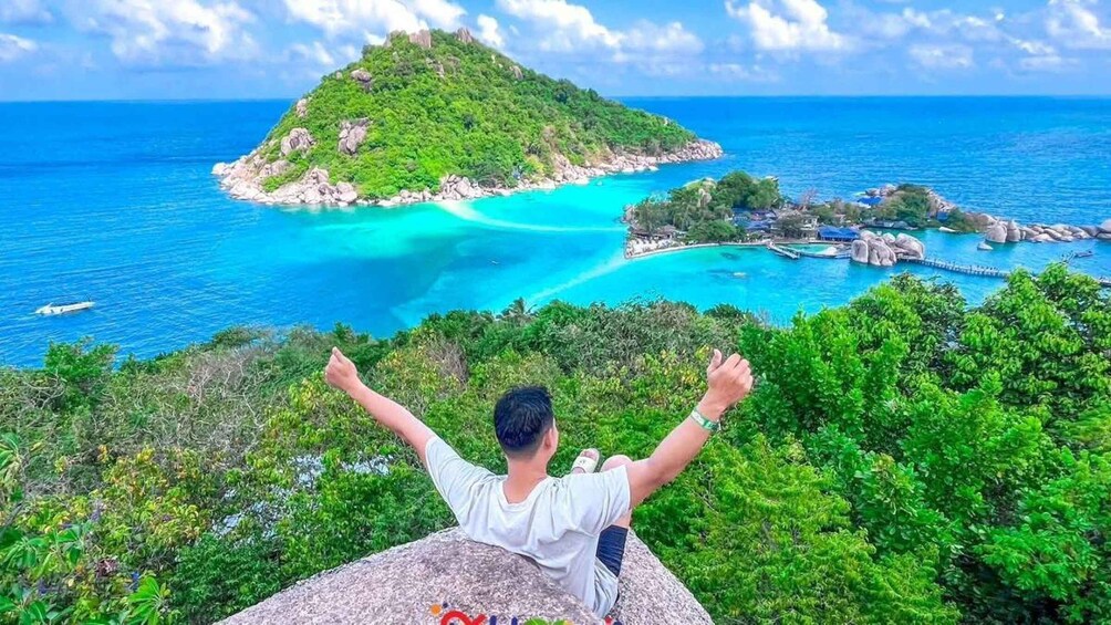 A person sitting on a rock with arms raised, overlooking a turquoise bay with a small island and clear blue waters. 1