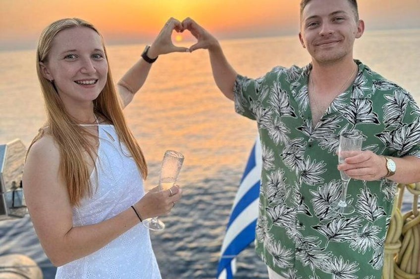 Two people making a heart shape with their hands, holding drinks, on a boat with a flag in the background. 4