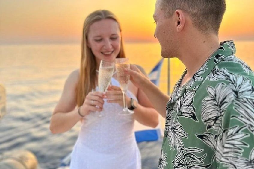 A person in a green shirt and a person in a white dress holding champagne glasses at sunset by the sea. 5
