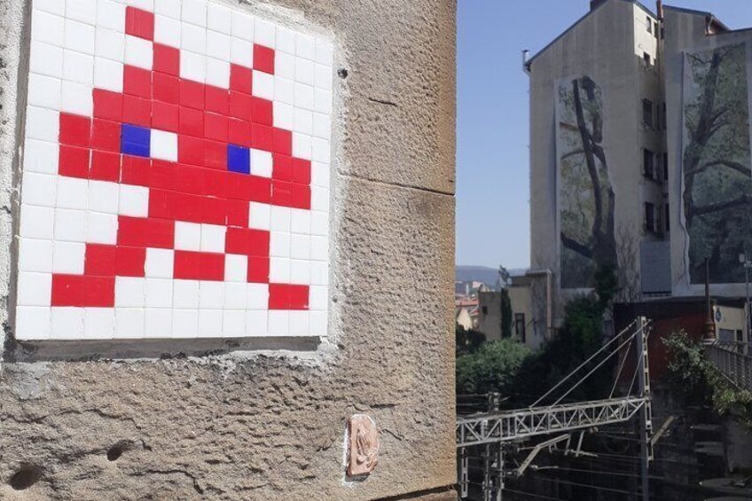 A pixelated red and white character on a wall, a building with tree designs, and scaffolding. 1