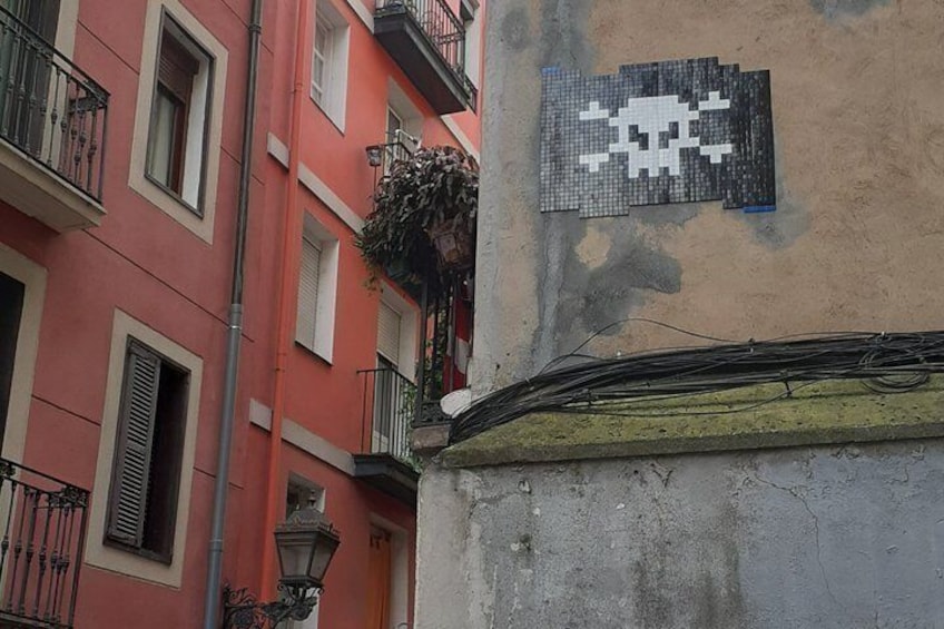 A building corner with a pixelated skull and crossbones sign, a red building, and a balcony with plants. 5
