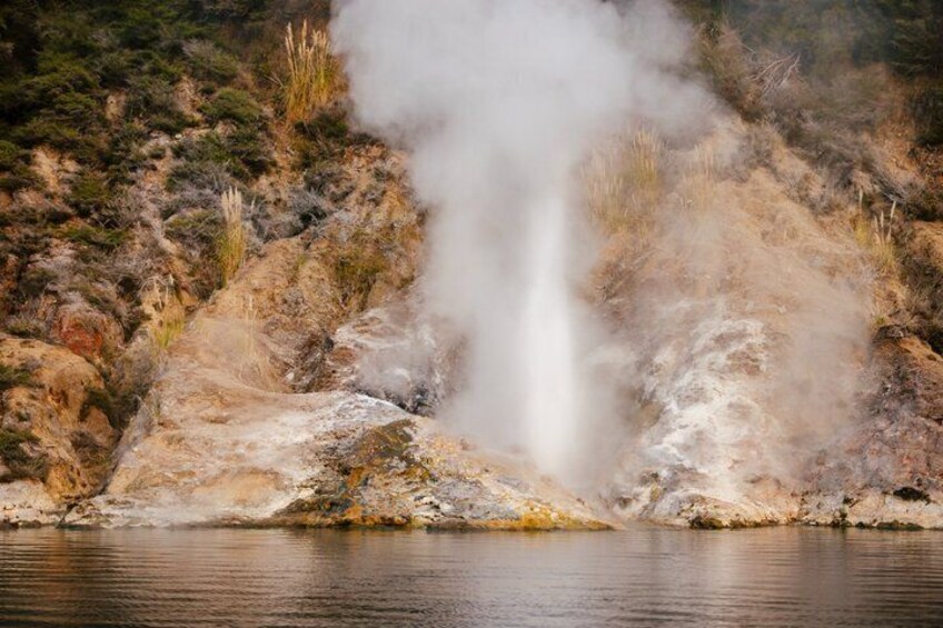 Un geyser con vapore che erutta, circondato da terreno roccioso e scarsa vegetazione. 2