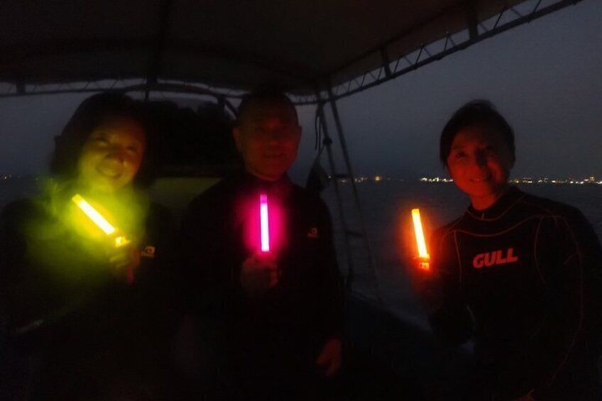Three people holding glowing sticks, with a boat and a cityscape in the background. 1