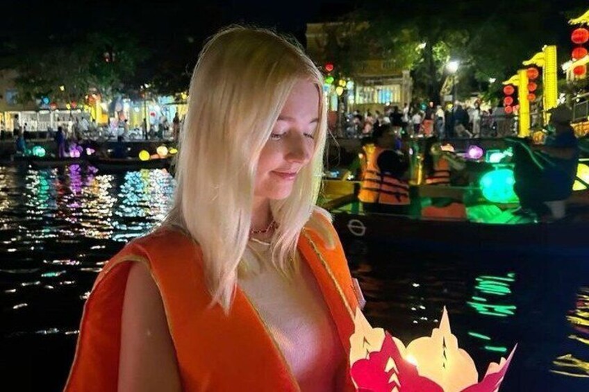 Una persona con un abito arancione in piedi vicino a un corpo d'acqua, con strutture illuminate sullo sfondo. 3