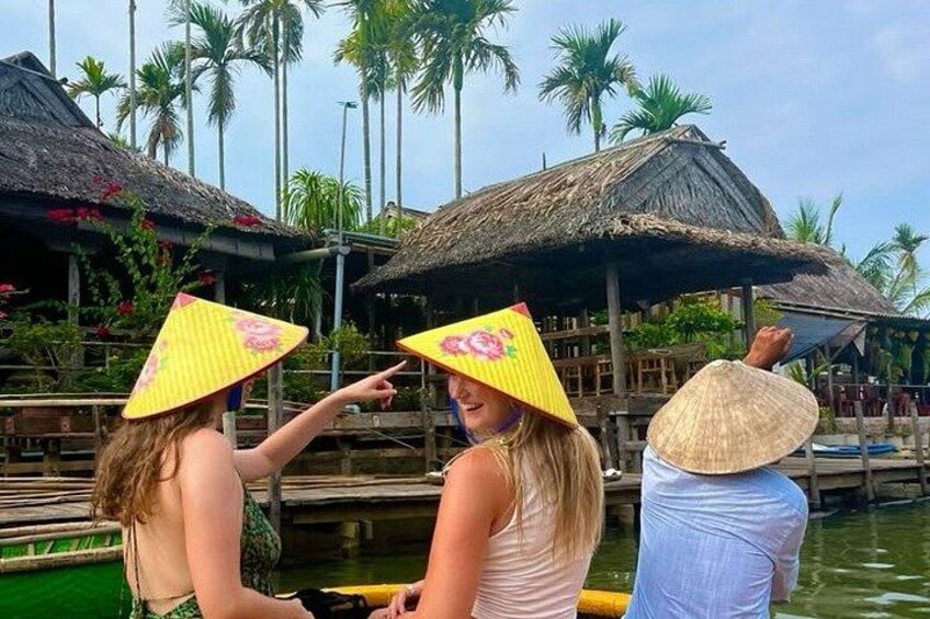 Dos personas en un bote, usando sombreros cónicos tradicionales, con estructuras de techo de paja y palmeras al fondo. 3