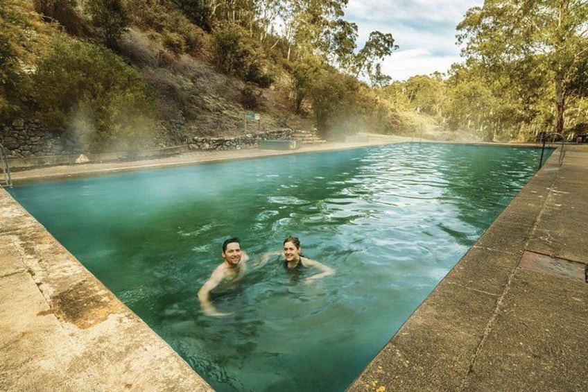 A natural hot spring with two people in the water, surrounded by trees and a stone wall. 5