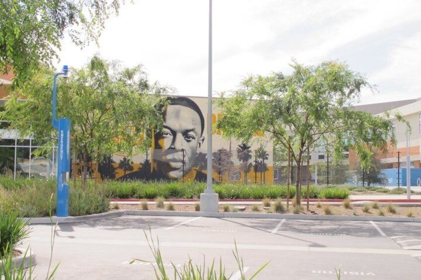 Un mural con el rostro de un hombre en un edificio, un farol azul y un estacionamiento con árboles. 2